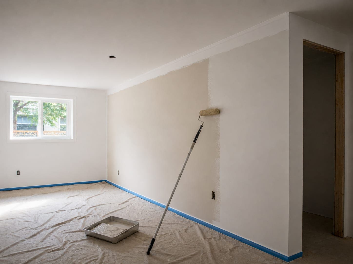 Interior wall being rolled with off-white paint over primer, painter's tape along the baseboard and drop cloths covering the floor